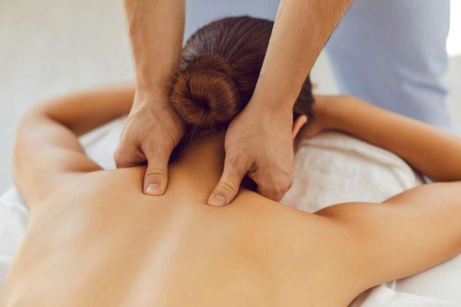 Gros plan d'un massage de la nuque, vu de dos, où les pouces d'un praticien exercent une pression ciblée sur les cervicales d'une femme aux cheveux bruns attachés en chignon.