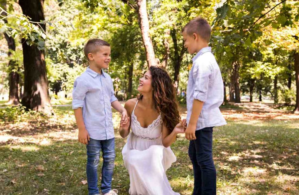 Mariée souriante vêtue de blanc discutant avec deux garçons dans un parc, illustrant comment impliquer les enfants dans le mariage.